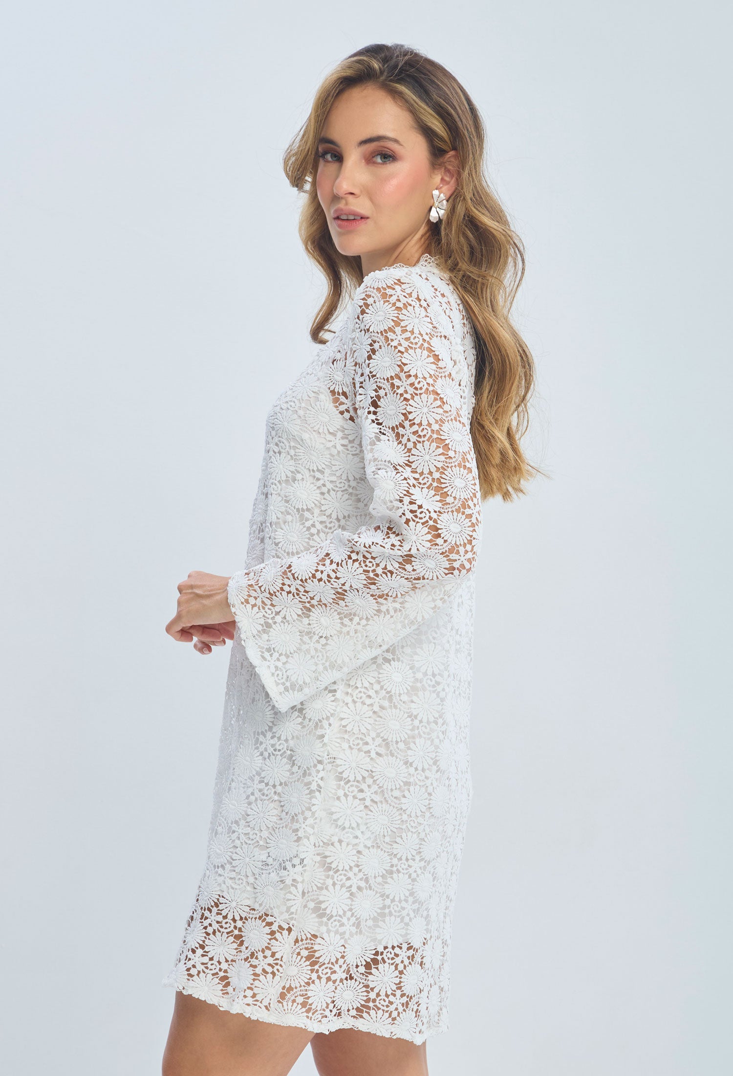 Woman in white floral lace dress with long sleeves, posing against a plain background