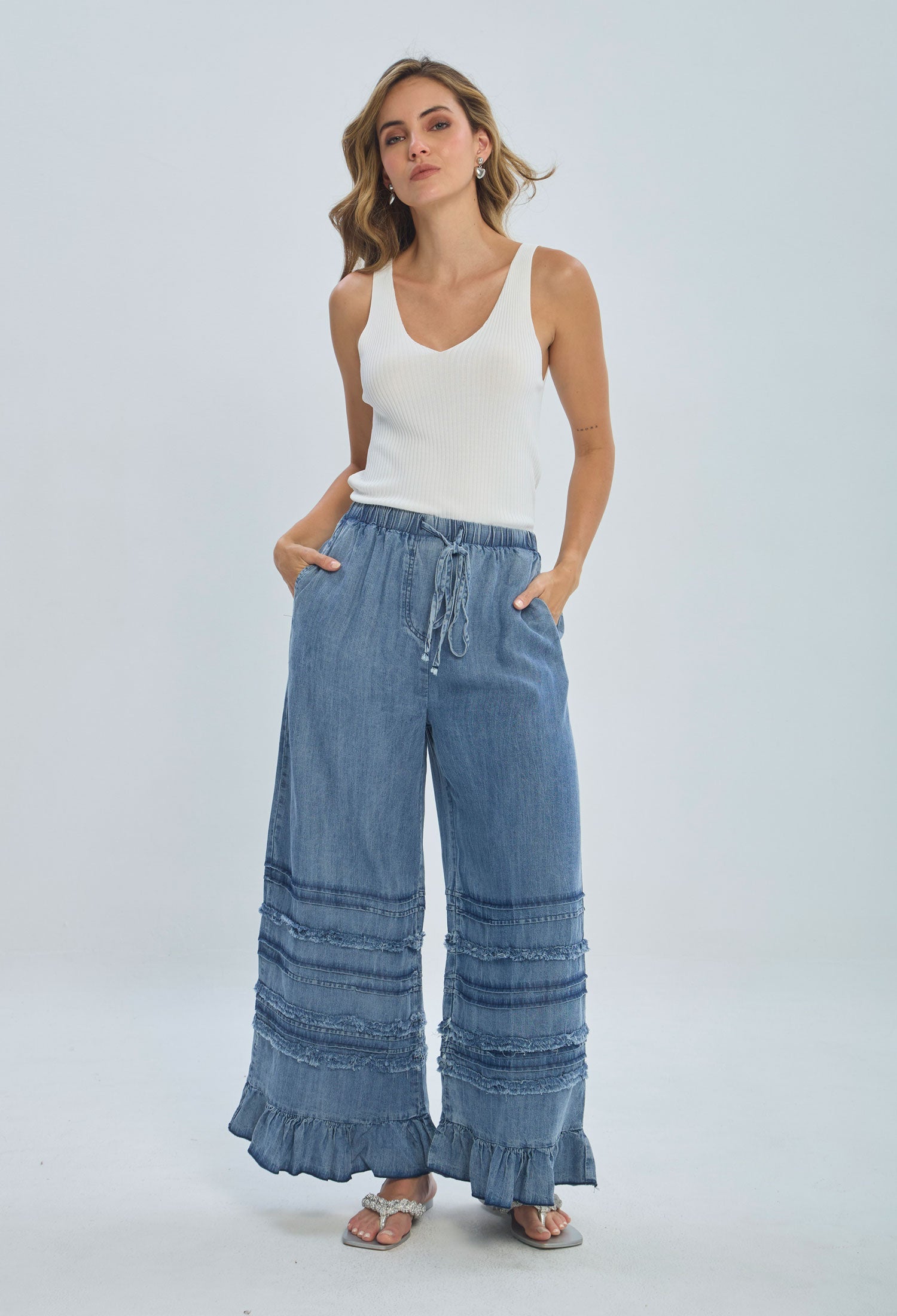 Woman wearing a white ribbed tank top and wide-leg denim pants with ruffle hem and lace trim.
