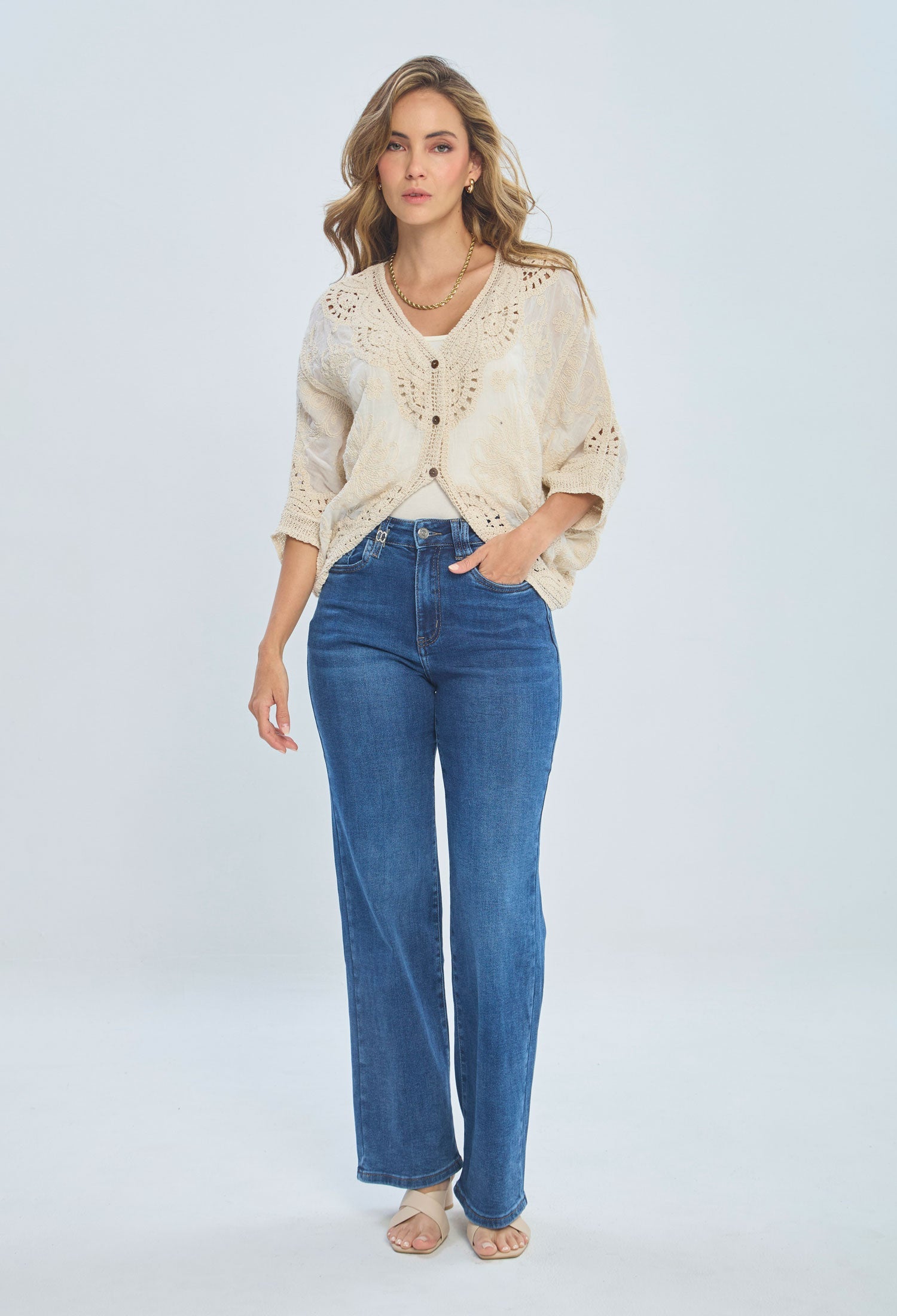 Woman wearing beige knit cardigan, high-waisted blue jeans, and nude sandals on white background