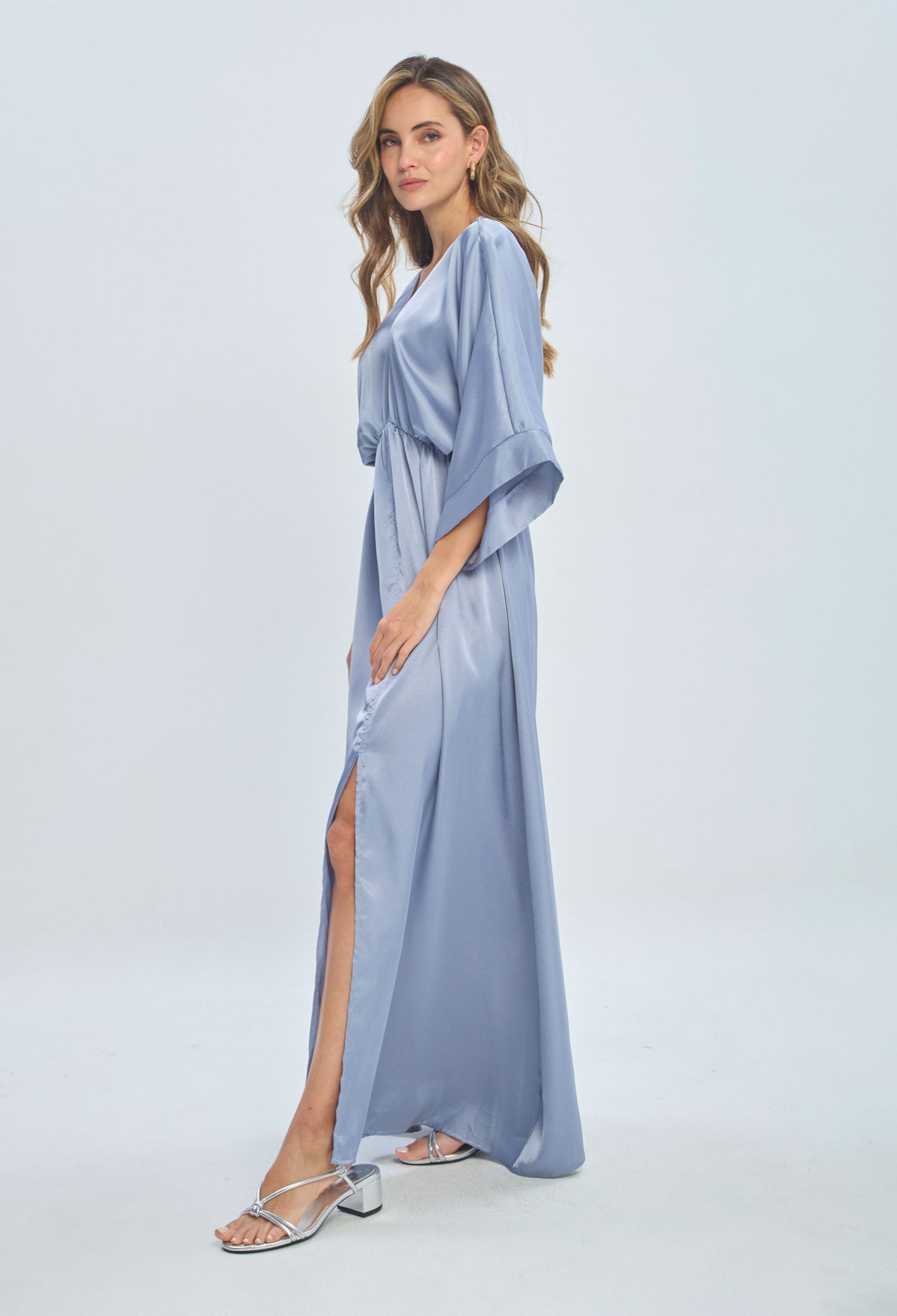 Woman in a light blue satin maxi dress with kimono sleeves and side slit, silver sandals