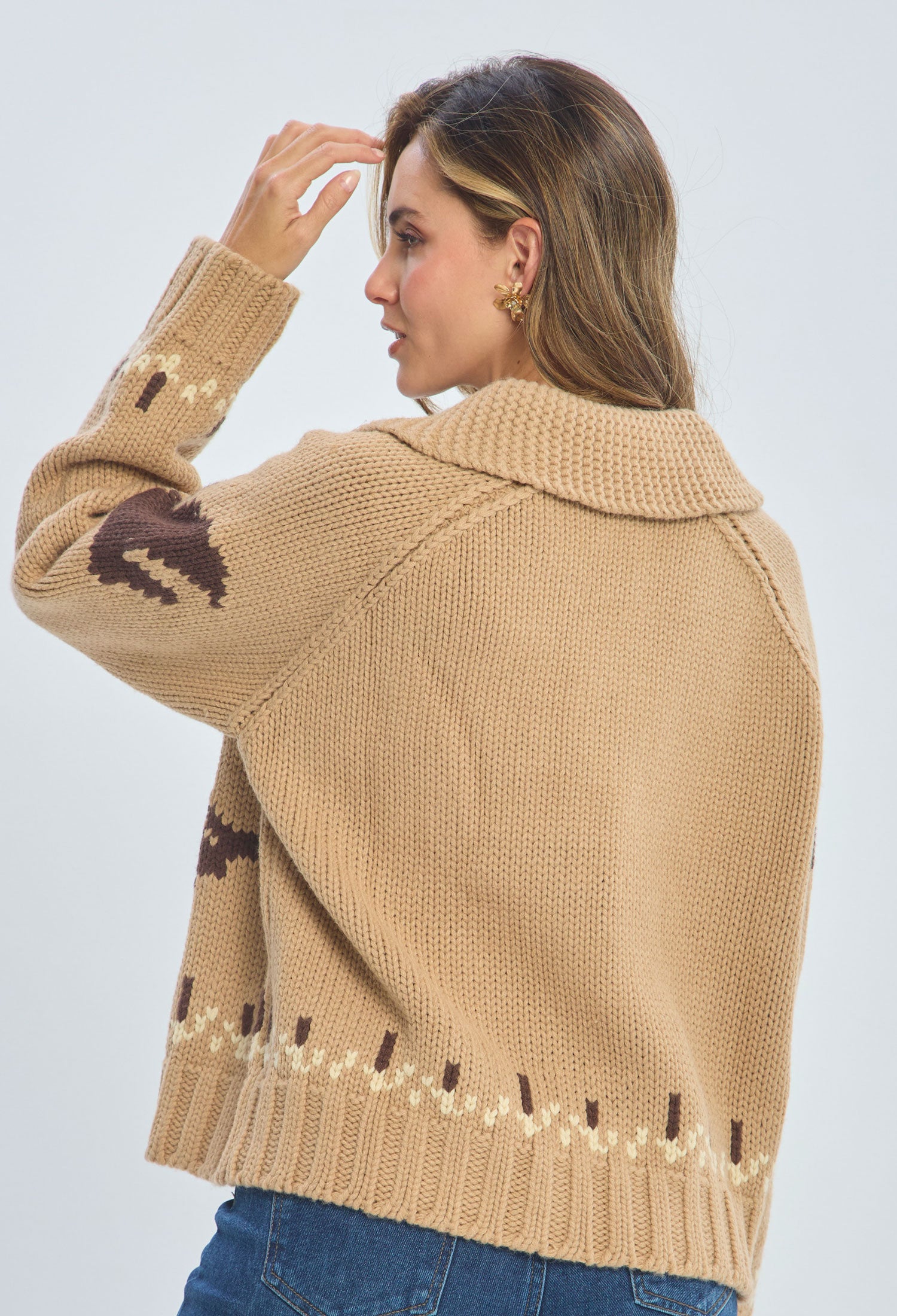Woman wearing a beige chunky knit sweater with brown patterns and blue jeans, back view